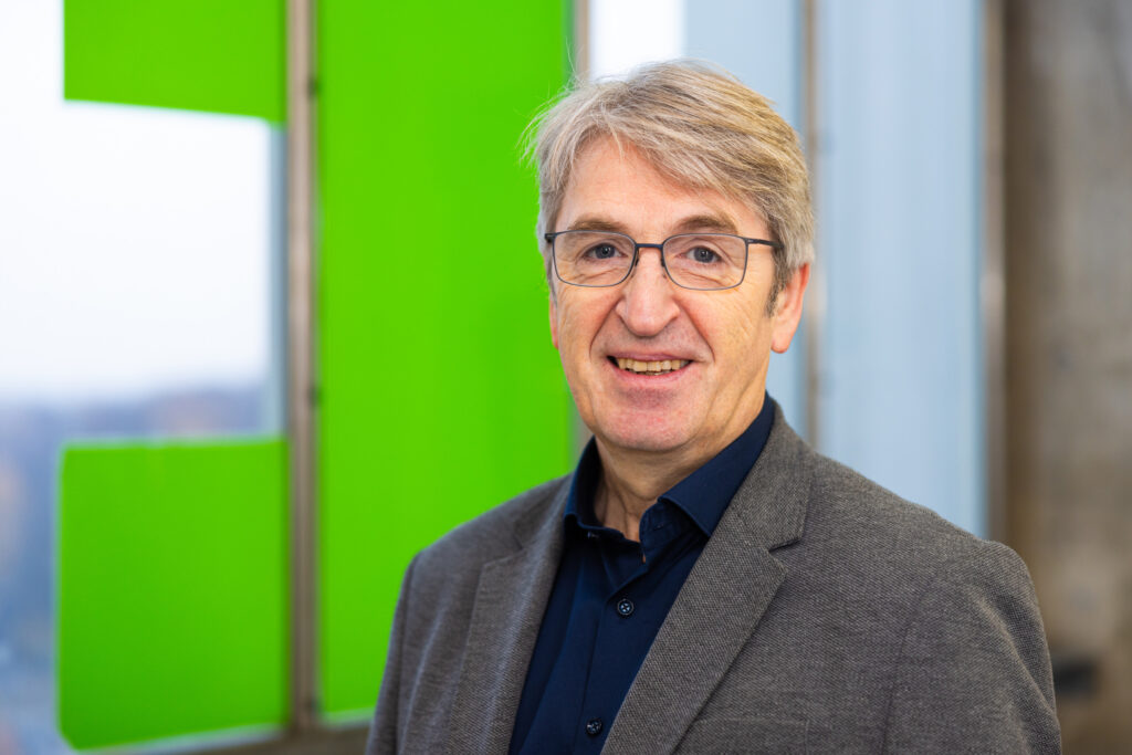 Porträt von Professor Dr. Ralf Vogel. Er hat eine Brille, trägt ein dunkles Hemd und ein graues Sakko und steht in einem modernen Innenraum vor einem unscharfen, grün-weißen Hintergrund.