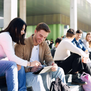 Auf der Treppe vor X-Gebäude setzten mehrere Studierende und sie schauen auf ein Tablet zu zweit.