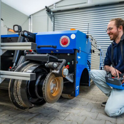 Eine Person kniet lächelnd auf dem Boden und hält ein Steuerungsgerät in der Hand, neben ihr ein blaues Spezialfahrzeug.