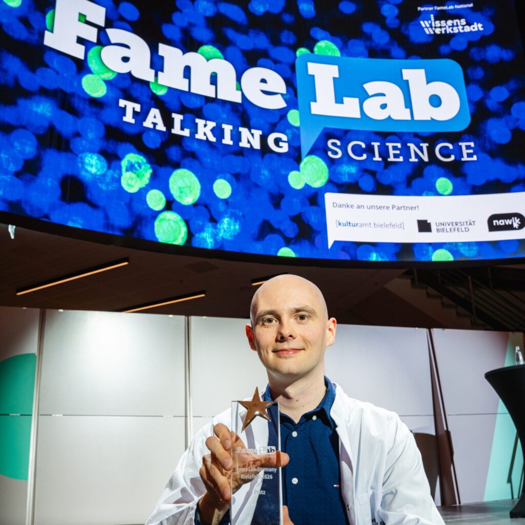 Person in Laborkittel ssitzt auf Bühne, im Hintergrund große Projektion "FameLab - Talking Science"