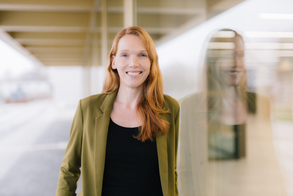 Professorin Dr. Marie I. Kaiser steht unter dem Vordach eines Gebäudes, rechts im Fenster eine Spiegelung von ihr.