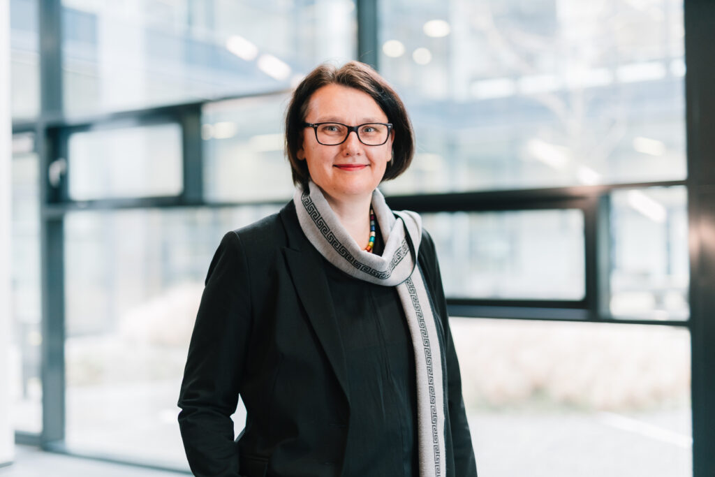 Professor Dr Britta Wrede, with dark hair and glasses, stands in front of large, bright window panels indoors. She is wearing a black outfit with a light-coloured scarf and is smiling slightly at the camera.
