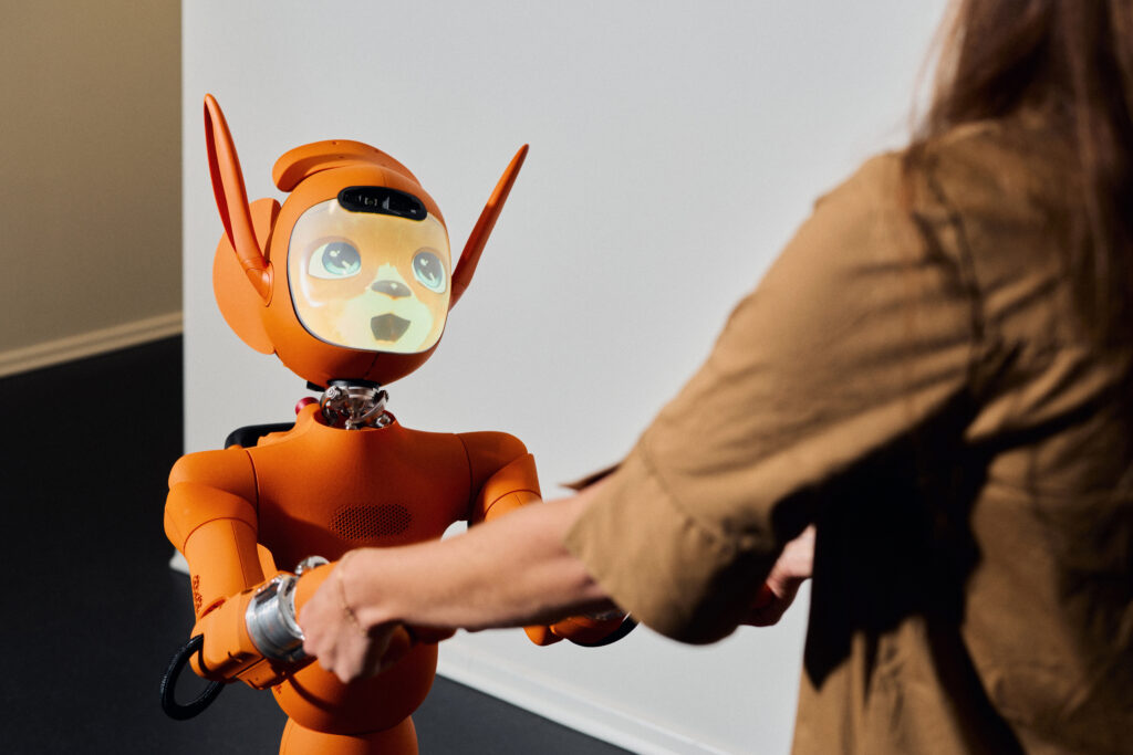 An orange robot with an animated face is holding the hands of a person standing opposite it.