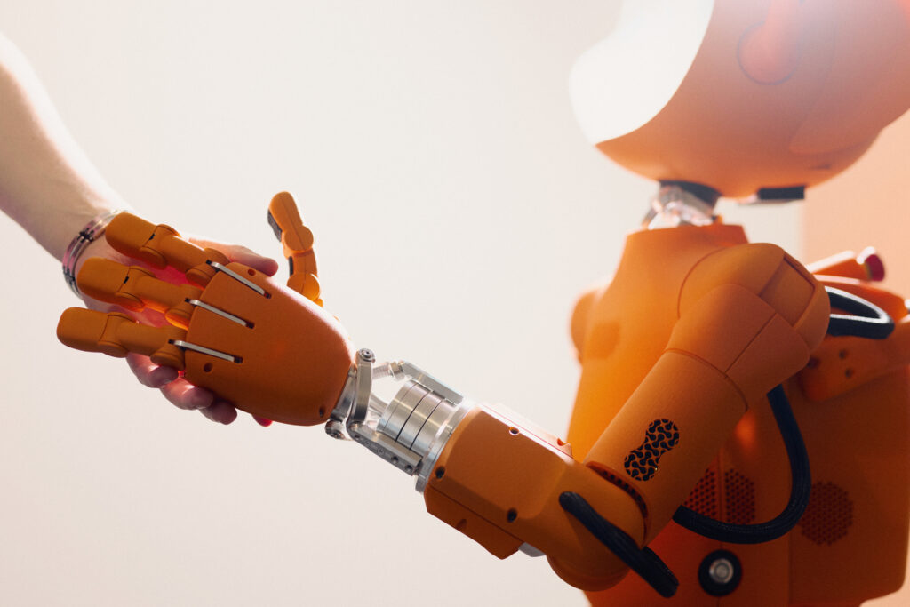 A human hand and an orange robotic hand are touching; on the right side of the image, the robot’s upper body is visible.
