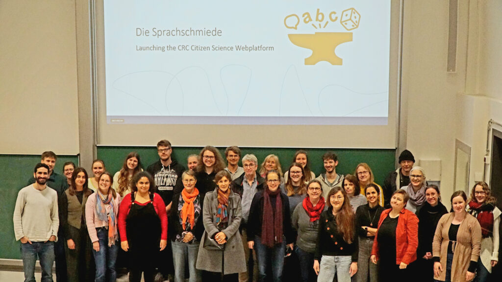 Gruppe steht vor Kreidetafeln in Hörsaal, oberhalb von ihr eine Leinwand mit Schriftzug "Die Sprachschmiede" und einem Logo