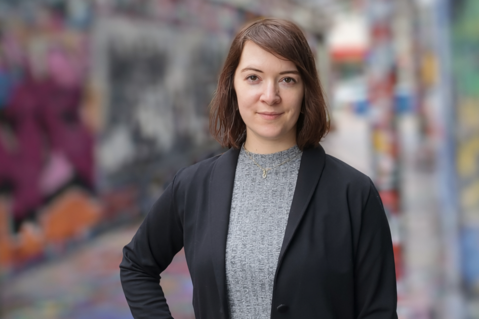 Dr. Kerstin Ostermann steht in einer schmalen, mit farbenreichen Graffiti bemalten Gasse. Sie trägt eine schwarze Jacke über einem grauen Oberteil und einen hellen, plissierten Rock. Eine Hand hat sie in die Hüfte gestützt, während sie direkt in die Kamera blickt.