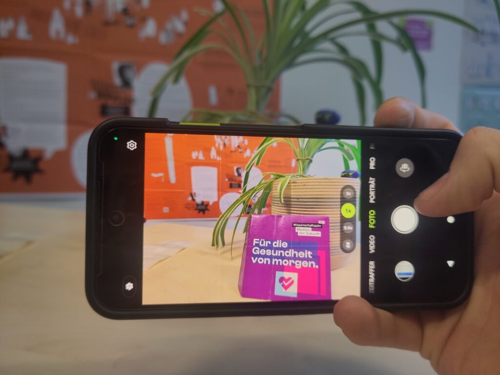Nahaufnahme einer Hand, die ein Smartphone im Querformat hält. Auf dem Display ist die Kamera-App aktiv und fokussiert eine lila Broschüre („Wissenschaftsjahr – Medizin der Zukunft“). Im Hintergrund ist eine Grünlilie in einem Topf und eine orangefarbene Infowand zu sehen.