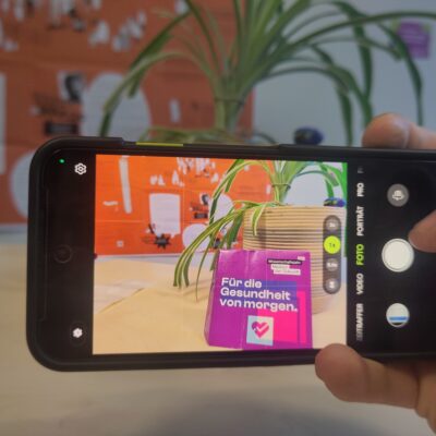 Nahaufnahme einer Hand, die ein Smartphone im Querformat hält. Auf dem Display ist die Kamera-App aktiv und fokussiert eine lila Broschüre („Wissenschaftsjahr – Medizin der Zukunft“). Im Hintergrund ist eine Grünlilie in einem Topf und eine orangefarbene Infowand zu sehen.