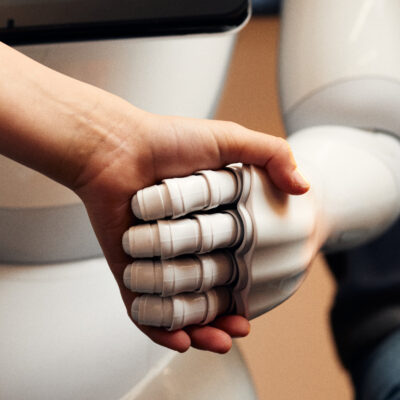 Nahaufnahme einer menschlichen Hand, die die Finger einer weißen Roboterhand hält. Beide Hände sind seitlich im Bild zu sehen, der Hintergrund ist unscharf.