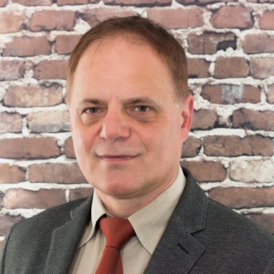 Porträt eines Mannes mit braunem Haar. Er trägt ein graues Sakko, ein helles Hemd und eine rote Krawatte. Sein Gesichtsausdruck ist neutral bis leicht freundlich. Im Hintergrund ist eine unscharfe Backsteinwand zu sehen.
