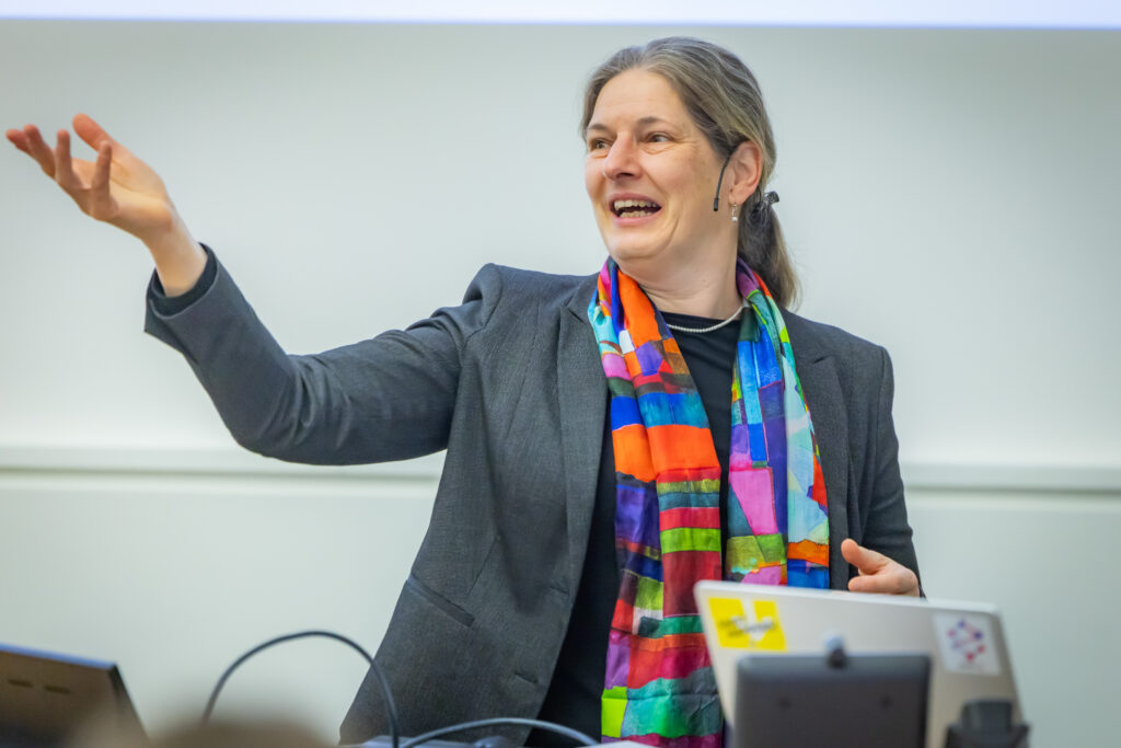 Frau in einem Vortragssaal, die zur Seite zeigt und lächelnd spricht. Sie trägt einen grauen Blazer und einen farbenfrohen, geometrisch gemusterten Schal. Vor ihr stehen ein Laptop und Präsentationstechnik, im Hintergrund eine helle, schlichte Wand.