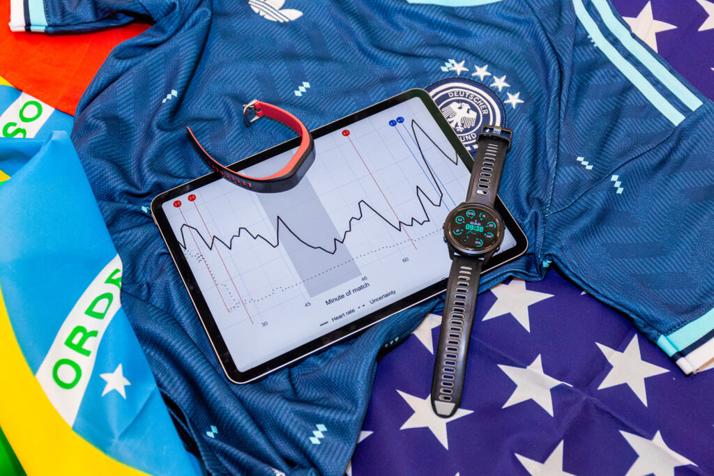 Auf mehreren Länderflaggen und einem deutschen Fußballtrikot liegen ein Tablet mit einer Herzfrequenzkurve, eine Garmin-Smartwatch und ein Fitness-Tracker.