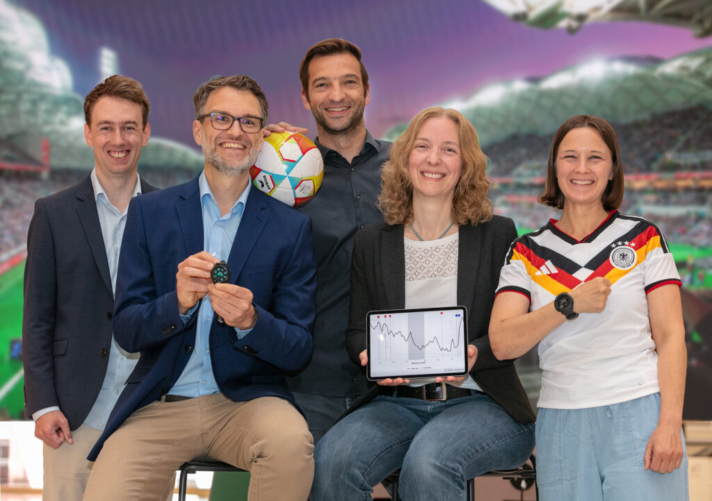 Fünf lächelnde Personen posieren vor dem unscharfen Hintergrund eines Fußballstadions. Ein Mann im blauen Sakko hält eine Smartwatch, während der Mann hinter ihm einen bunten Fußball hält. Eine Frau präsentiert ein Tablet mit einem Liniendiagramm und die Frau ganz rechts trägt ein Trikot der deutschen Nationalmannschaft mit einer Sportuhr am Handgelenk.