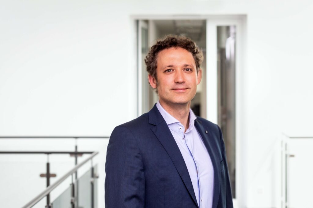 A man in a suit in a bright office corridor, smiling slightly at the camera.