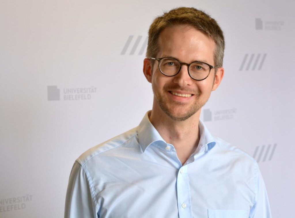 Ein lächelnder Mann mit Brille, kurzen braunen Haaren und Dreitagebart im Portrait. Er trägt ein hellblaues Hemd. Der Hintergrund ist weiß und mit dem Logo der Universität Bielefeld gemustert.