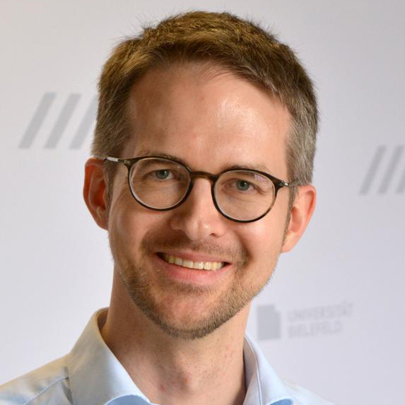 Ein lächelnder Mann mit Brille, kurzen braunen Haaren und Dreitagebart im Portrait. Er trägt ein hellblaues Hemd. Der Hintergrund ist weiß und mit dem Logo der Universität Bielefeld gemustert.