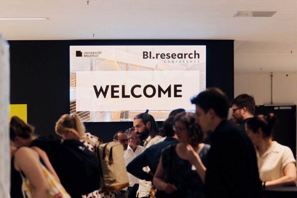 Mehrere Personen bewegen sich vor einem großen Bildschirm, der die Schriftzüge "Welcome" und "BI.research Conference" zeigt.