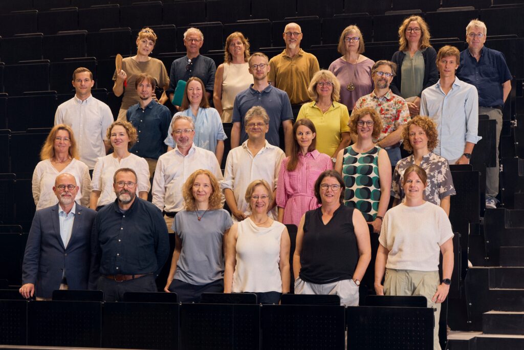 Gruppenfoto von 27 Frauen und Männern, die in mehreren Reihen in einem Hörsaal stehen und in die Kamera blicken.