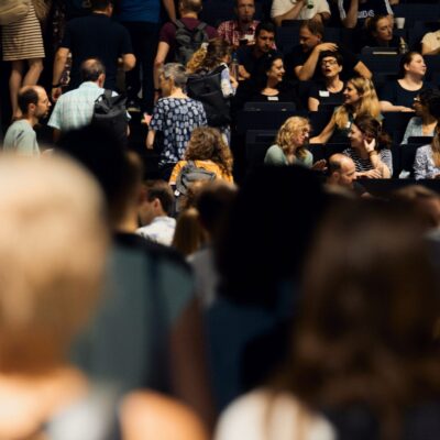 Blick in einen dicht besetzten Hörsaal aus der Perspektive des Eingangs oder Gangs. Im stark unscharfen Vordergrund sieht man die Hinterköpfe und Schultern von mehreren Personen, die in den Saal hineingehen oder dort stehen. Der Fokus liegt auf dem mittleren und hinteren Teil des Raums, wo zahlreiche Teilnehmende auf den ansteigenden Sitzreihen Platz genommen haben oder sich auf den Treppen bewegen. Viele der sitzenden Personen unterhalten sich angeregt in Kleingruppen miteinander oder blicken nach vorne.