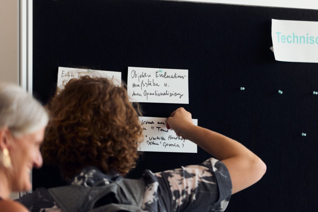 Nahaufnahme einer schwarzen Pinnwand, an die weiße Moderationskarten mit handschriftlichen Notizen geheftet sind. Im Fokus steht von hinten eine Person mit kurzen, braunen Locken, deren Arm angehoben ist, um eine Karte festzustecken. Links im Anschnitt ist das Profil einer lächelnden Frau mit grauen Haaren und einem großen Ohrring zu sehen.