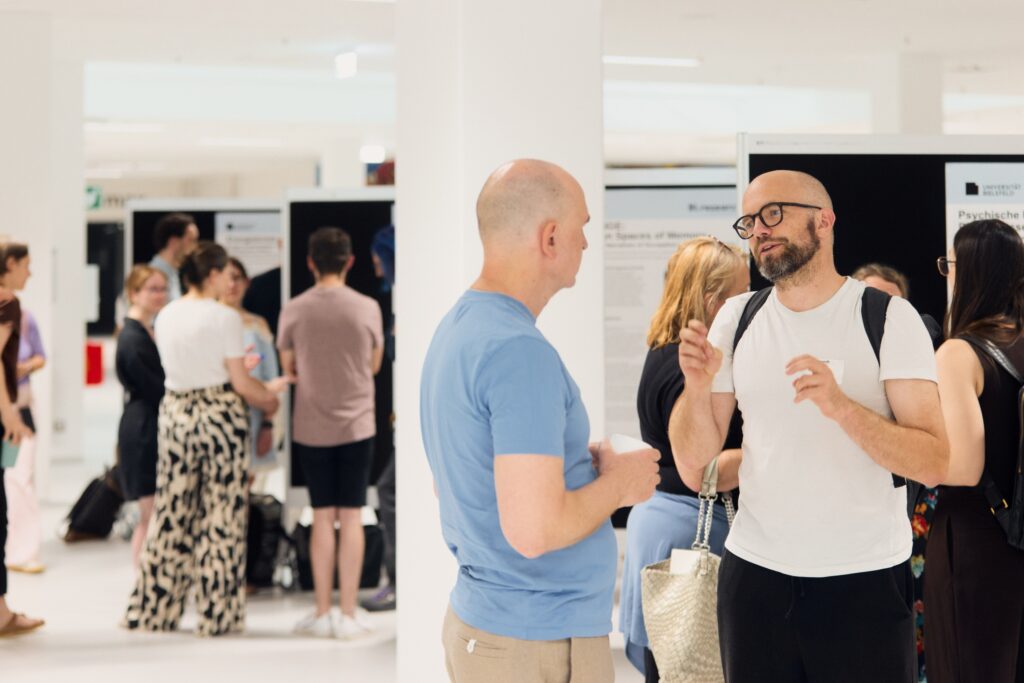 Zwei Männer im Gespräch bei einer Posterausstellung in einem hellen Ausstellungsraum. Im Hintergrund weitere Teilnehmende.