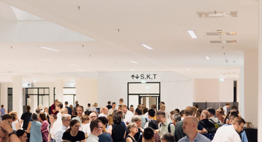 Große Gruppe von Menschen, die in der hellen, weitläufigen Halle des Hauptgebäudes der Universität Bielefeld dicht beieinander stehen. Die Teilnehmenden der BI.research Conference 2026 sind in Gespräche vertieft. Im Hintergrund sind weiße Wände mit Wegweisern zu sehen, die Decke ist hoch und mit integrierten Lichtleisten ausgestattet.