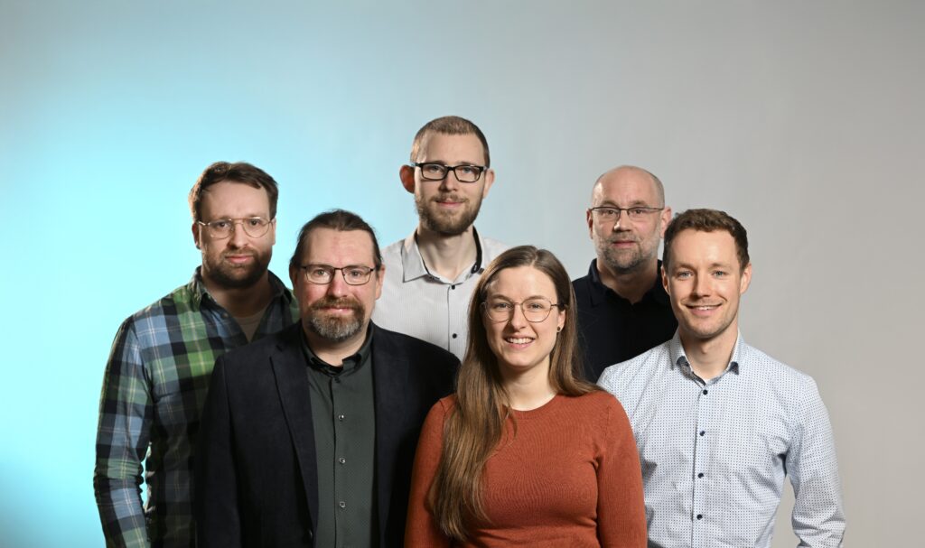 Fünf Personen vor einem neutralen Hintergrund, der links blau ausgeleuchtet ist