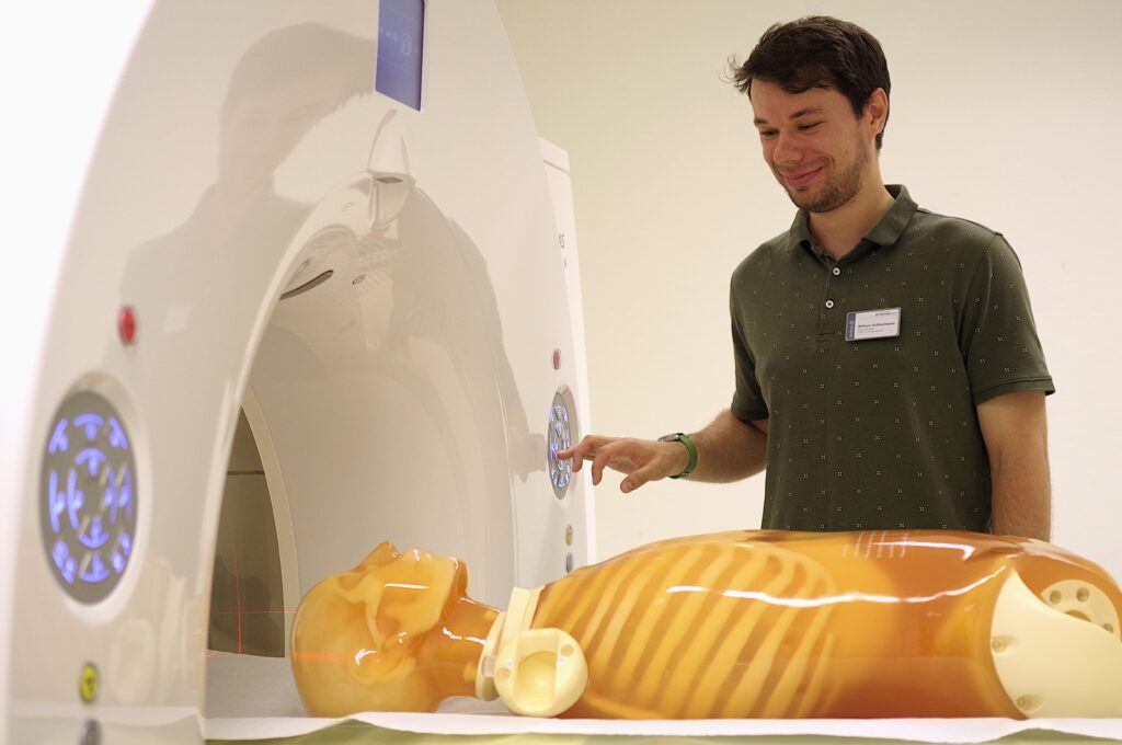 Ein Medizinphysiker bedient lächelnd ein CT-Gerät über ein Bedienfeld, während ein transparenter Phantom-Torso zur Optimierung der Bildgebung auf der Patientenliege liegt.