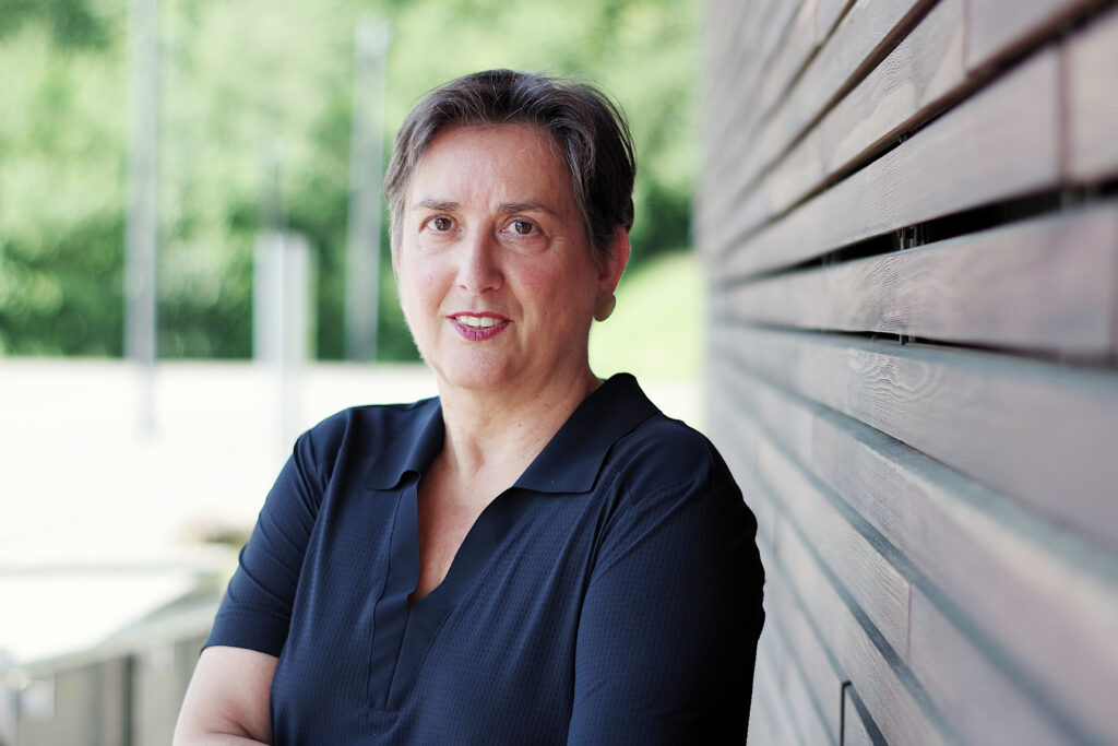 Eine Frau in einem blauen Hemd steht neben einer Holzfassade und lächelt in die Kamera.