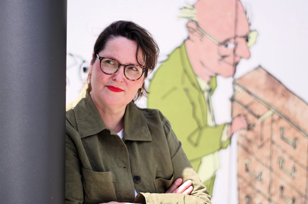 Eine Frau steht an eine Säule gelehnt; sie trägt eine Brille und eine grüne Jacke. Hinter ihr ist an der Wand die Zeichnung eines Mannes mit Brille zu sehen.