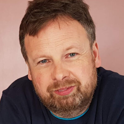 Nahaufnahme eines Mannes mittleren Alters mit kurzen, braunen Haaren, Bart und blauen Augen, der leicht lächelt. Er trägt einen dunkelblauen Pullover.