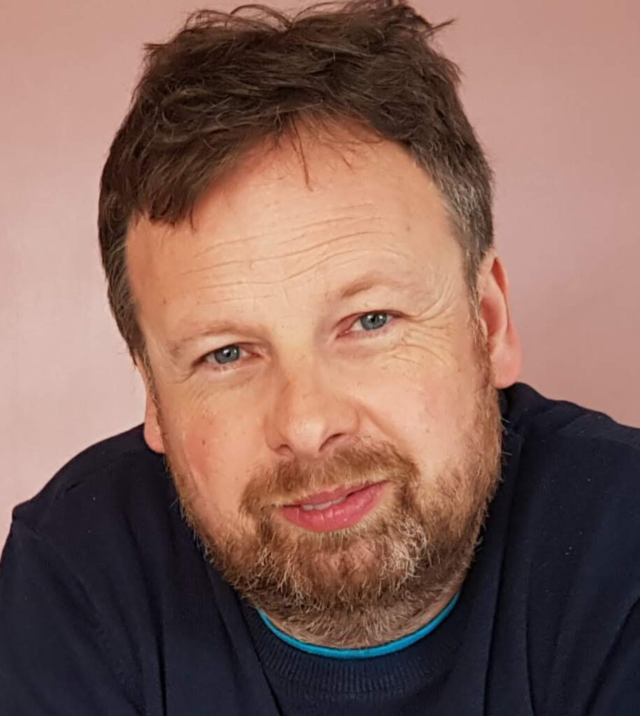 Close-up of a middle-aged man with short brown hair, a beard and blue eyes, smiling slightly. He is wearing a dark blue jumper.