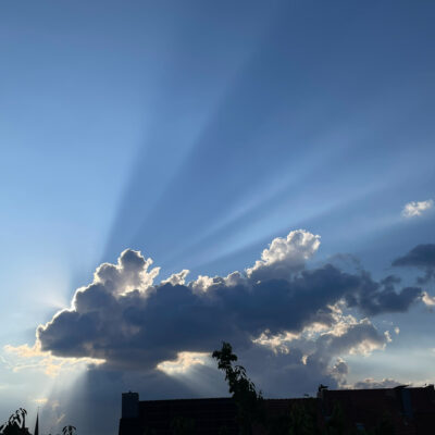 Himmel mit Sonnenstrahlen, die hinter einer dunklen Wolke hervorbrechen, über einer Silhouette von Dächern und Bäumen.