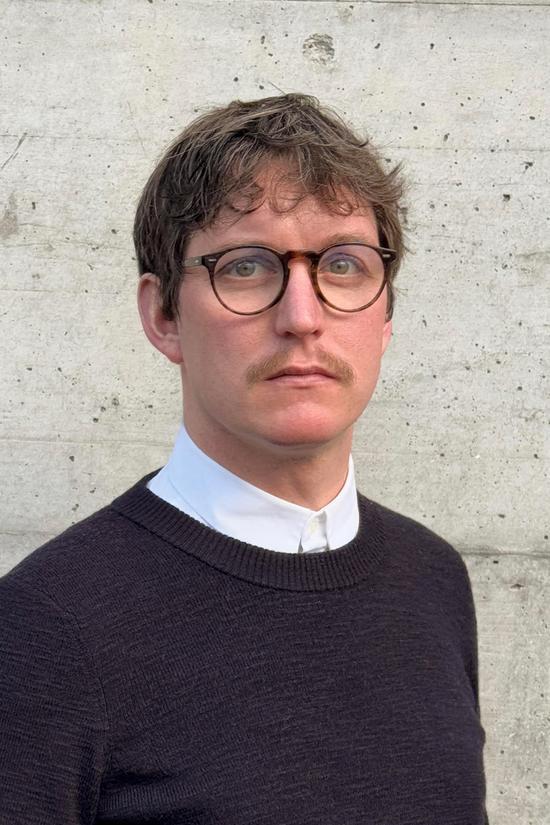 Porträtfoto eines Mannes mit Brille und Schnurrbart, der einen dunklen Pullover über einem weißen Hemd mit Kragen trägt, vor einer hellen Betonwand.