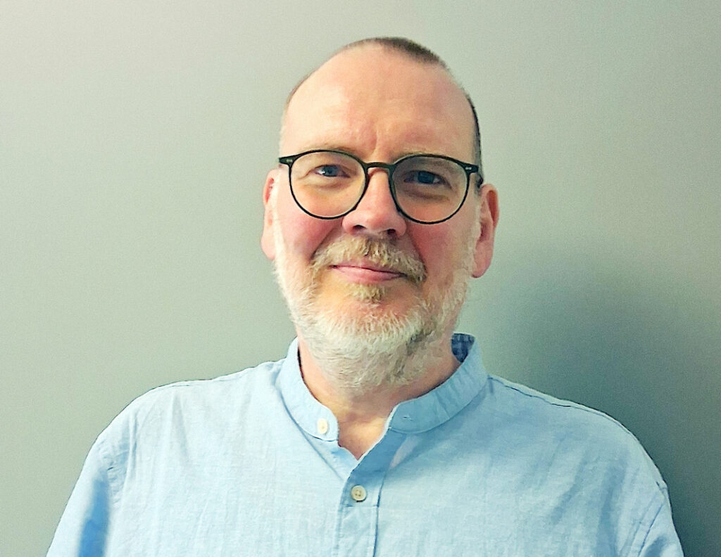 Porträtfoto eines Mannes mit Brille, grauem Bart und hellblauem Hemd vor einer einfarbigen grauen Wand.