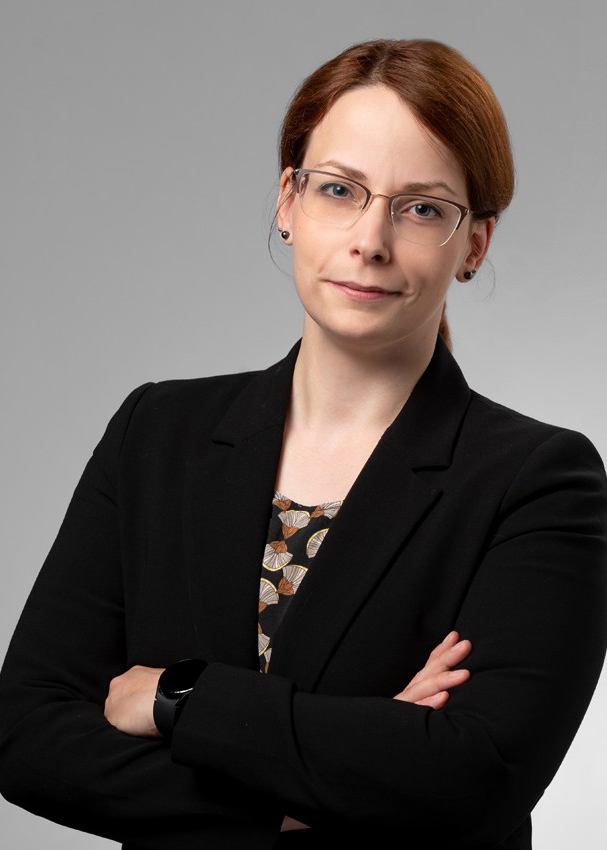 Porträtfoto einer Frau mit Brille und zurückgebundenem rötlich-braunem Haar, die einen schwarzen Blazer über einem gemusterten Oberteil trägt und die Arme verschränkt hat.