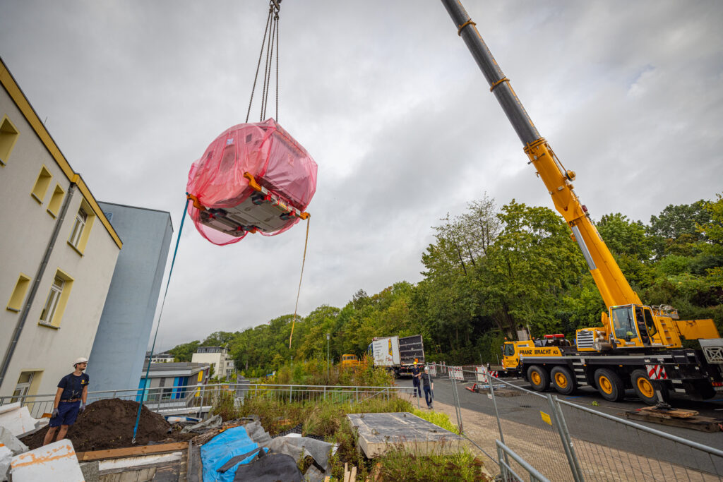 Gelber Mobilkran hebt ein mit rosa Folie umhülltes MRT-Gerät über eine Baustelle, im Hintergrund sind ein mehrstöckiges Gebäude und Bäume zu sehen.