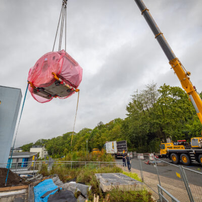 Gelber Mobilkran hebt ein mit rosa Folie umhülltes MRT-Gerät über eine Baustelle, im Hintergrund sind ein mehrstöckiges Gebäude und Bäume zu sehen.