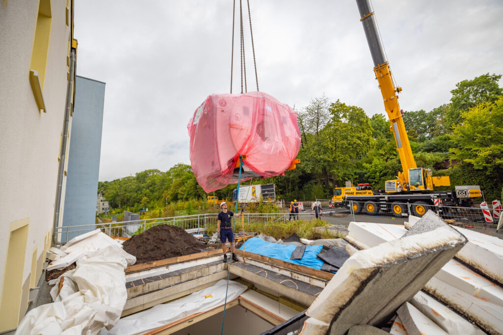 Gelber Mobilkran hebt ein mit rosa Folie umhülltes MRT-Gerät über eine Baustelle, im Hintergrund sind ein mehrstöckiges Gebäude und Bäume zu sehen.