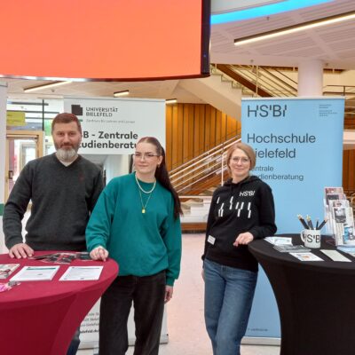 Auf dem Bild sind drei Personen zu sehen: Links ist ein Mann und rechts neben ihm sind zwei Frauen in der Informationsstelle am Tisch