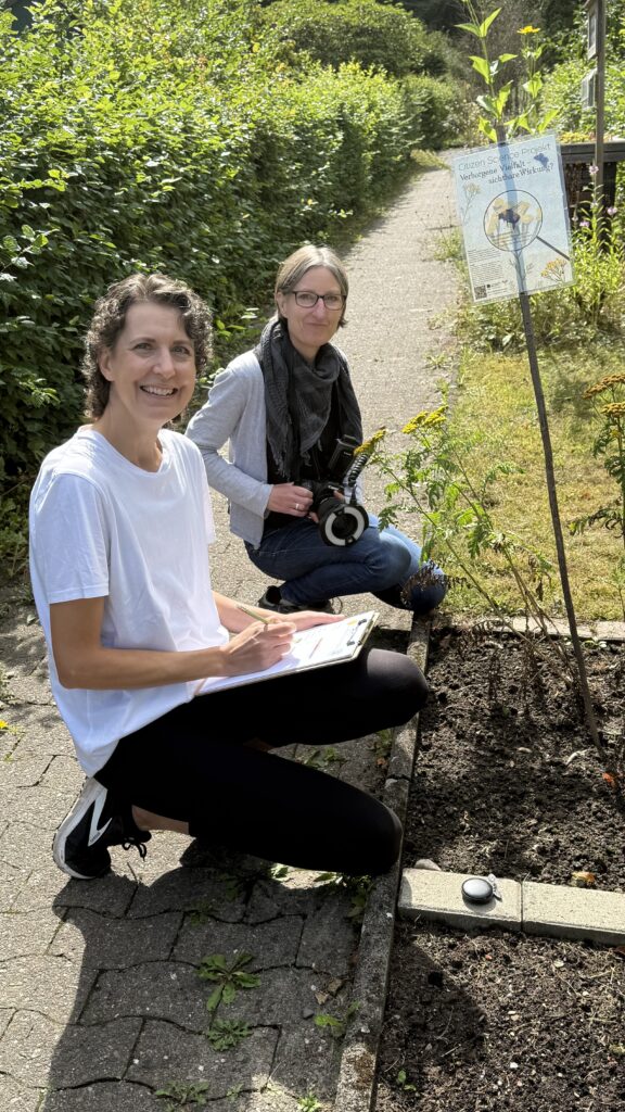Zwei Frauen knien vor einem Beet mit Pflanzen und machen sich Notizen. Sie lachen in die Kamera.
