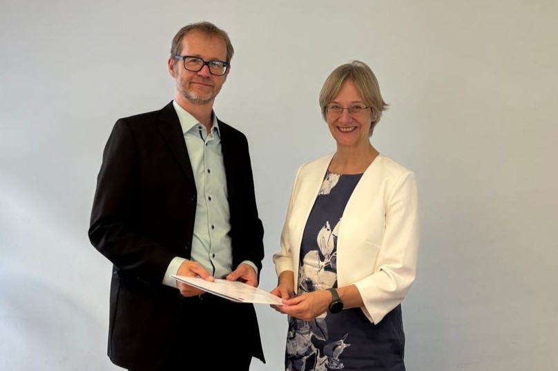 Foto eines Mannes im Anzug, der neben einer Frau steht, die ein Kleid trägt. Beide tragen eine Brille. Beide zusammen halten das unterzeichnete Framework Agreement in ihren Händen.
