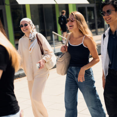 Foto: Drei Studierende laufen im Sonnenschein über den Campus der Universität Bielefeld. Im Hintergrund sieht man das X-Gebäude.